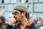 Man wearing sunglasses and beanie blowing a whistle in an urban setting with crowd.