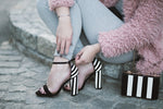 Anonymous woman in stylish shoes with striped handbag sitting on pavement and fastening thin straps on trendy high heeled sandals on daytime
